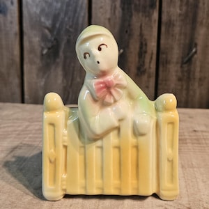 Puede incluir: Una maceta de cerámica vintage con la forma de un bebé con gorro inclinado sobre una cuna amarilla. La figura del bebé tiene un lazo rosa y está pintada en suaves colores pastel. El artículo es decorativo.