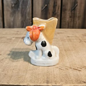 Pode incluir: Um pequeno vaso de cerâmica vintage em forma de vaca sentada. A vaca é branca com manchas pretas e um laço laranja. A parte do vaso é de cor amarelo claro.