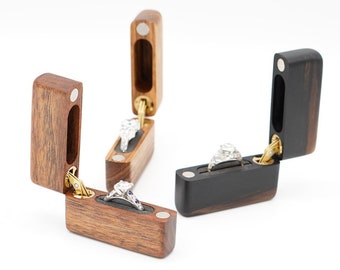 May include: Three wooden ring boxes with hinged lids. Two boxes are made of light brown wood and one is made of dark brown wood. Each box has a ring inside.