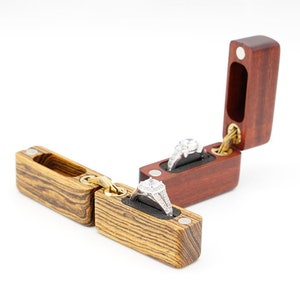 Puede incluir: Dos cajas de madera para anillos con tapas con bisagras. Las cajas están hechas de diferentes tipos de madera, una con un acabado marrón rojizo y la otra con una veta de madera marrón claro. Cada caja tiene un interior de terciopelo y se muestra un anillo en cada caja.