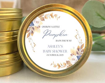 May include: A gold-colored tin with a white label featuring the text "OH BOY! A LITTLE Pumpkin IS ON THE WAY! ASHLEY'S BABY SHOWER OCTOBER 26, 2025". The label has floral and leaf accents in shades of blue, brown, and white. The tin is round.