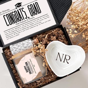 May include: A black gift box filled with brown shredded paper. The box contains a white heart-shaped dish with the initials "NR" in black, a clear glass bottle with a cork top, and a brown candle with a pink and white label that says "Happy Graduation". The box also has a card with the text "Congrats, Grad" and a message for Stacey.