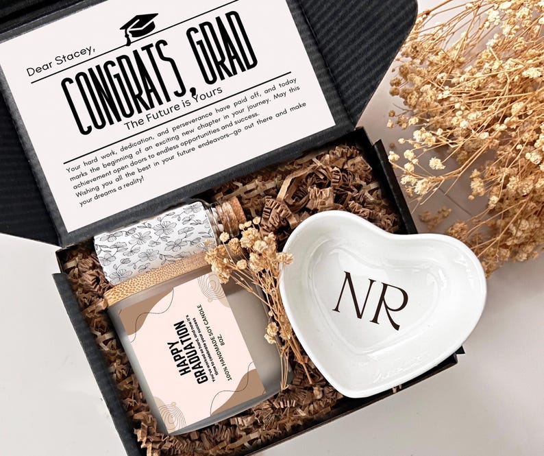 May include: A black gift box filled with brown shredded paper. The box contains a white heart-shaped dish with the initials "NR" in black, a clear glass bottle with a cork top, and a brown candle with a pink and white label that says "Happy Graduation". The box also has a card with the text "Congrats, Grad" and a message for Stacey.