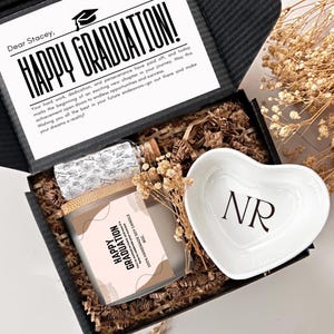 May include: A black gift box filled with brown packing peanuts. The box contains a white heart-shaped dish with the initials "NR" printed on it, a small glass bottle with a cork stopper, and a soy candle with a label that reads "Happy Graduation".
