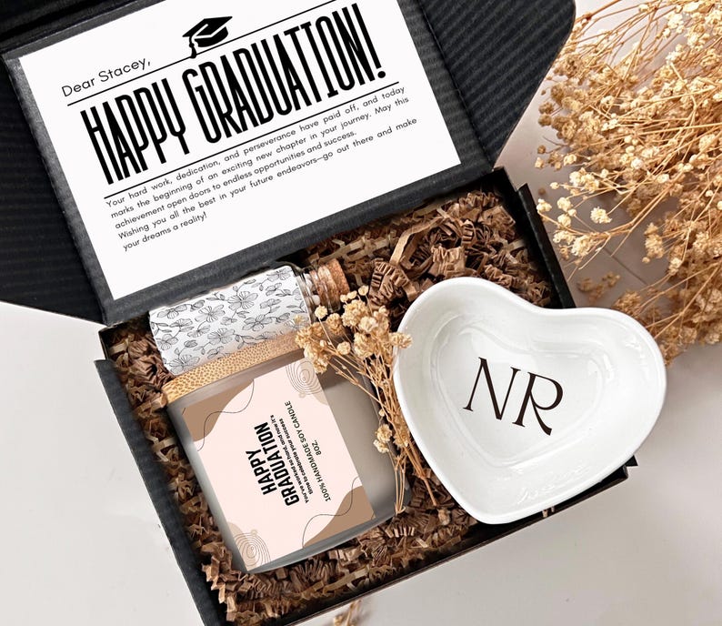 May include: A black gift box filled with brown packing peanuts. The box contains a white heart-shaped dish with the initials "NR" printed on it, a small glass bottle with a cork stopper, and a soy candle with a label that reads "Happy Graduation".