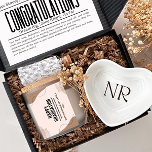 May include: A black gift box with a white card that reads "Congratulations, Dream Big, Shine Bright, Go Far". The box contains a soy candle with a pink and white label that reads "Happy Graduation", a small heart-shaped dish with the initials "NR" in black, and a clear glass bottle with a cork stopper.