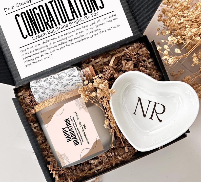 May include: A black gift box with a white card that reads "Congratulations, Dream Big, Shine Bright, Go Far". The box contains a soy candle with a pink and white label that reads "Happy Graduation", a small heart-shaped dish with the initials "NR" in black, and a clear glass bottle with a cork stopper.