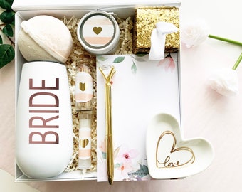 May include: A white gift box filled with a variety of items, including a white tumbler with the word "BRIDE" in rose gold lettering, a bath bomb, a small jar of lotion, a heart-shaped dish with the word "love" written in gold, a gold pen, and a notepad with a floral design.