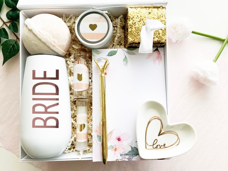 May include: A white gift box filled with a variety of items, including a white tumbler with the word "BRIDE" in rose gold lettering, a bath bomb, a small jar of lotion, a heart-shaped dish with the word "love" written in gold, a gold pen, and a notepad with a floral design.