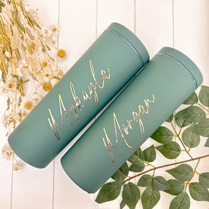 May include: Two teal-colored insulated tumblers with gold lettering. One tumbler is labeled "Mkayla" and the other "Morgan". The tumblers are cylindrical with a matching lid. The background includes dried flowers and greenery.