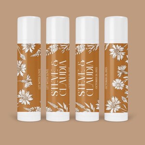May include: Four lip balm tubes with white caps and brown labels. The labels feature white floral designs and text that reads "LIT WITH LOVE", "STEVE & CLAUDIA", and "OCTOBER 26, 2025". The background is a solid brown color.