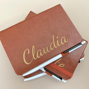 May include: Two brown leather journals with gold lettering. The top journal has the name "Claudia" written on the front. Both journals have black and white pens attached to them with orange elastic bands.