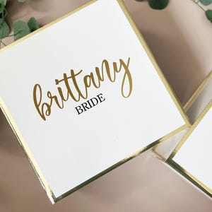 May include: White square gift box with gold trim and the name "Brittany" in gold script, with "BRIDE" in a smaller, black font. The box is part of a set, with another box visible in the background.