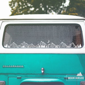 May include: A teal blue Volkswagen van with a white mountain range silhouette decal on the rear window. The decal features a crescent moon and stars. The van has the word "VOLKSWAGEN" in white letters on the back.