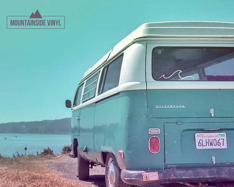 Puede incluir: Una furgoneta Volkswagen azul verdosa con techo blanco y una pegatina de ola blanca en la ventana trasera. La furgoneta est&aacute; aparcada en un camino de tierra con una masa de agua en el fondo. El texto "MOUNTAINSIDE VINYL" est&aacute; en la esquina superior izquierda de la imagen. La matr&iacute;cula dice "6LHW067".