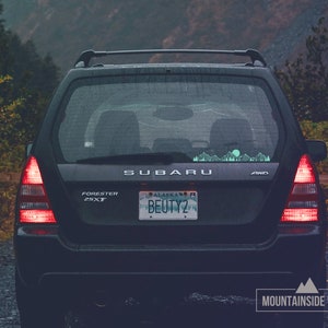 May include: A black Subaru Forester 2.5XT with a license plate that reads "ALASKA BEUTY2" and a mountain range decal on the back window. The car is parked on a gravel road in a forest.