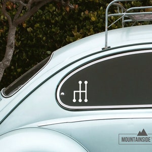 Op de afbeelding: Een witte vinyl sticker van een pookversnellingschema op de achterruit van een lichtblauwe vintage auto. De auto heeft een dakrek en de sticker is geplaatst op de ruit achter de bestuurdersstoel.