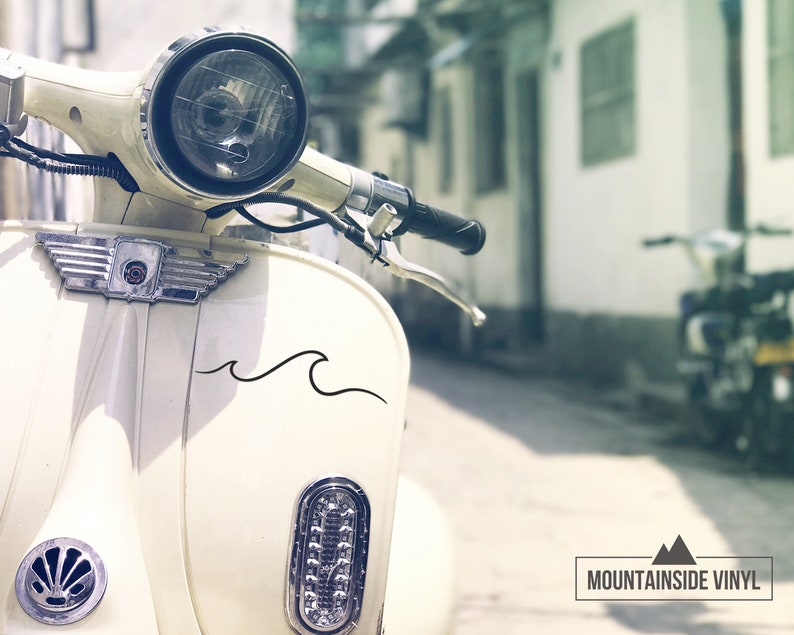 Puede incluir: Un scooter vintage blanco con una calcoman&iacute;a de onda negra en el lateral. El scooter est&aacute; estacionado en un callej&oacute;n estrecho con un edificio en el fondo. El scooter tiene un faro cromado y un manillar cromado.