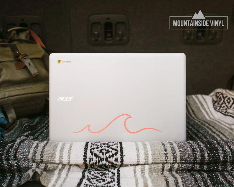 Puede incluir: Un ordenador port&aacute;til blanco con una pegatina de onda rosa en la tapa. El port&aacute;til est&aacute; sentado en una manta a rayas en la parte trasera de un coche. El port&aacute;til tiene la palabra "acer" y "chrome" en la tapa.