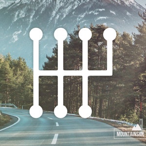 May include: White vinyl sticker of a 5-speed manual transmission gear shift pattern. The sticker is on a road with a mountain range in the background.