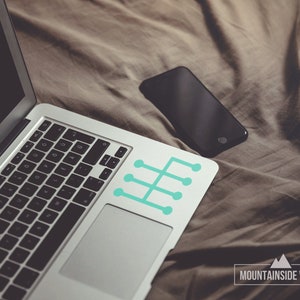 Op de afbeelding: Een zilveren laptop met een zwart toetsenbord en een turquoise blauwe sticker met een versnellingsbakpatroon op de rechterbovenhoek van het touchpad. De laptop ligt op een bruine deken met een zwarte smartphone ernaast. De tekst "MOUNTAINSIDE VINYL" staat onderaan de afbeelding.