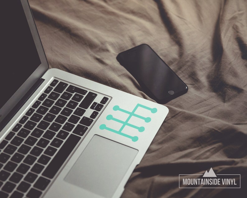 Op de afbeelding: Een zilveren laptop met een zwart toetsenbord en een turquoise blauwe sticker met een versnellingsbakpatroon op de rechterbovenhoek van het touchpad. De laptop ligt op een bruine deken met een zwarte smartphone ernaast. De tekst "MOUNTAINSIDE VINYL" staat onderaan de afbeelding.