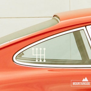 Op de afbeelding: Een rode auto met een witte 5-versnellingsbak-versnellingsbak-sticker op de achterruit. De sticker is een populair ontwerp voor autoliefhebbers. De auto staat geparkeerd voor een witte muur. De tekst "MOUNTAINSIDE VINYL" is zichtbaar in de rechterbenedenhoek van de afbeelding.