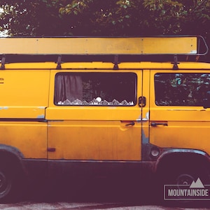 May include: A yellow van with a roof rack and a mountain range sticker on the side window. The van is parked in front of a green bush.