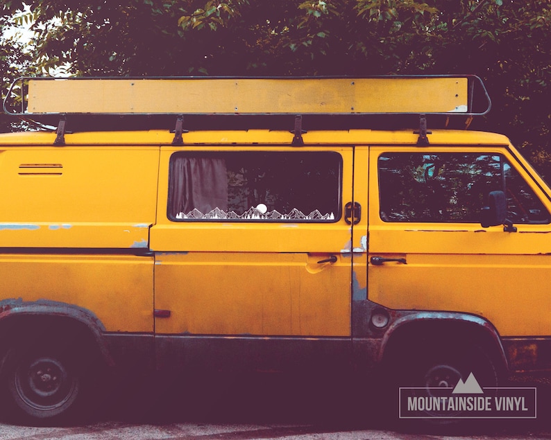 May include: A yellow van with a roof rack and a mountain range sticker on the side window. The van is parked in front of a green bush.