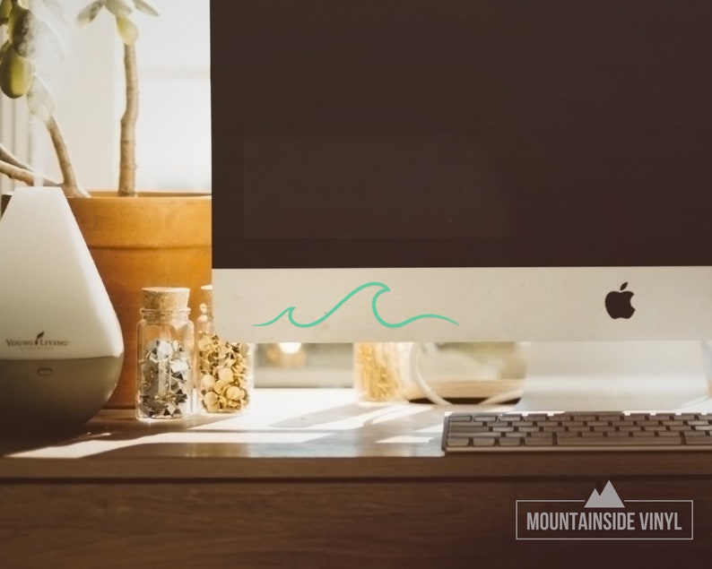 Puede incluir: Un monitor de computadora blanco con una pegatina de onda turquesa en el borde inferior. El monitor est&aacute; sobre un escritorio de madera con un teclado delante. El texto "MOUNTAINSIDE VINYL" es visible en la esquina inferior derecha de la imagen.