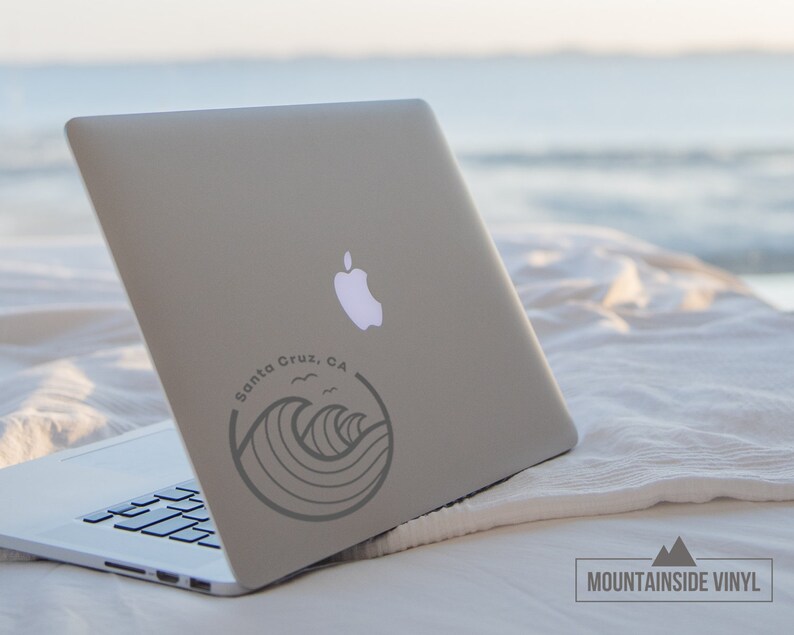 May include: A laptop computer with a sticker on the back that says "Santa Cruz, CA" with a graphic of waves and seagulls. The laptop is on a white blanket with a beach in the background.