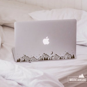 May include: A laptop computer with a mountain range silhouette decal on the lid. The decal is black and features a crescent moon and stars. The laptop is on a white bedspread.