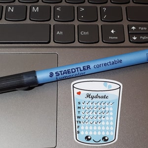 Op de afbeelding: Een blauwe en witte sticker met een cartoon glas water met de tekst "Hydrate" en vinkjes voor elke dag van de week. Een rood hartje bevindt zich bovenaan het glas. Een blauwe en zwarte Staedtler Lumocolor corrigeerbare pen ligt op een laptop.