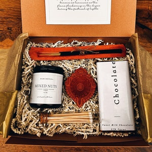 May include: A gift box filled with Diwali themed items, including a candle, incense sticks, a small clay diya, and a bar of chocolate. The box is lined with shredded paper and the lid has a card with the message "Happy Diwali. May every aspect of your life become as luminous as the flame flickering on the diyas during the festival of lights."