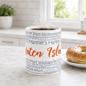 May include: A white ceramic mug filled with dark coffee, featuring the orange text Staten Island and various blue neighborhood names including Port Richmond, Mariner's Harbor, Fort Wadsworth, Springville, and Stapleton printed on the exterior.