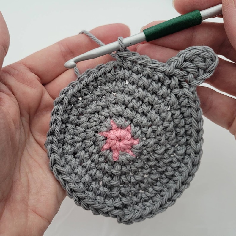 May include: A gray crocheted circle with a pink star in the center. The circle has a crocheted ear on the right side.