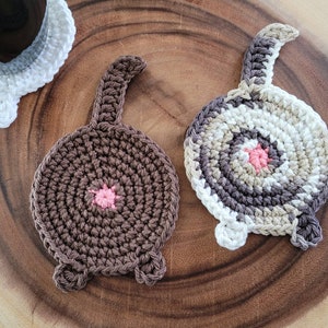 May include: Two crocheted cat-shaped coasters. One is brown with a pink nose and the other is white with a brown, beige, and pink nose.