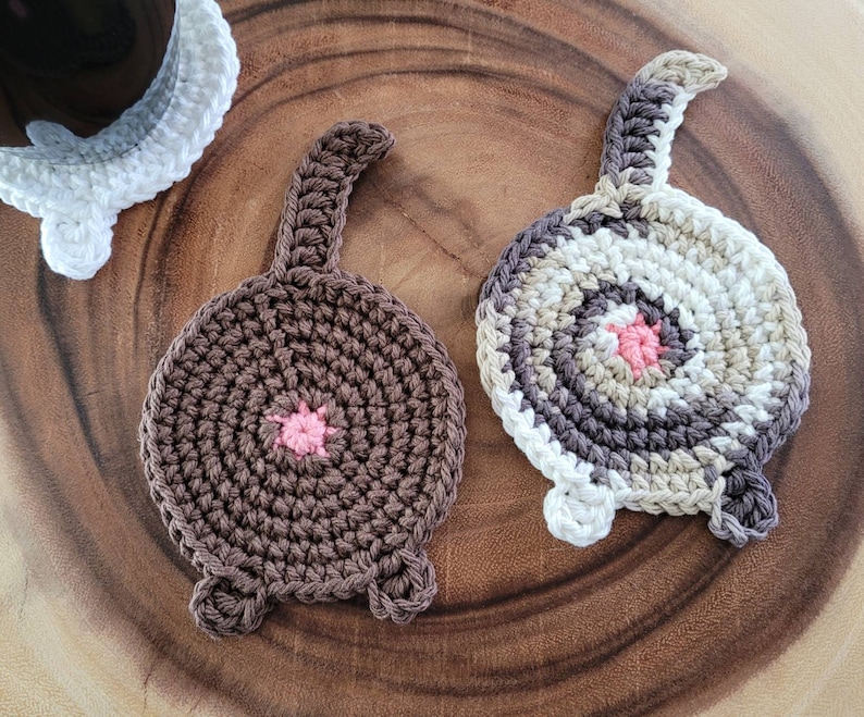 May include: Two crocheted cat-shaped coasters. One is brown with a pink nose and the other is white with a brown, beige, and pink nose.