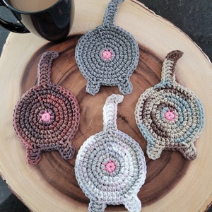 May include: Four crocheted cat butt coasters in different colors. The coasters are made with a variety of brown, gray, and white yarn. Each coaster has a pink circle in the center.