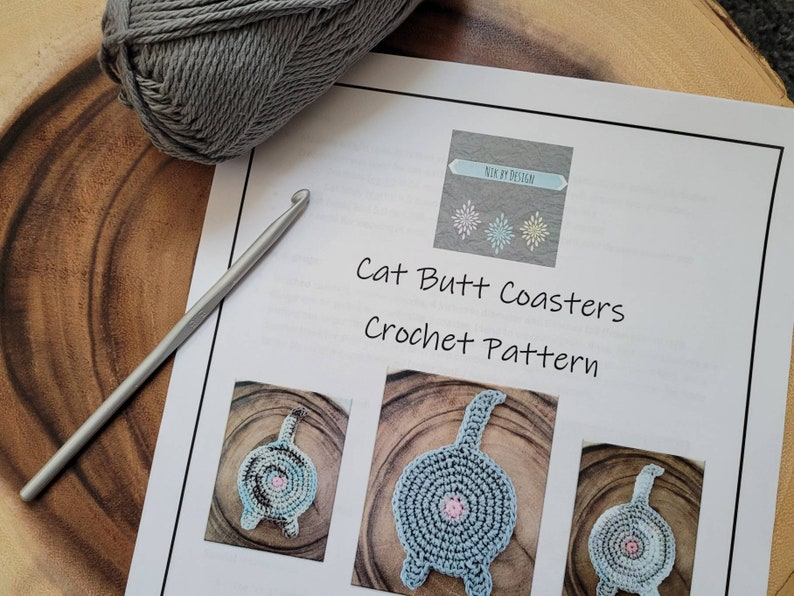 May include: A crochet pattern for cat butt coasters. The pattern includes instructions and images of the finished product. The coasters are gray with a pink center and a tail.