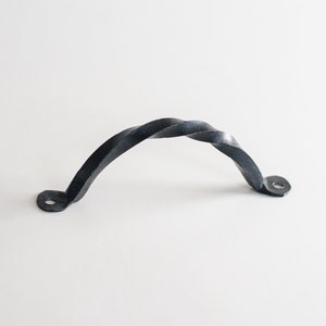 May include: A hand-forged, black iron handle with a twisted design. The handle is arched with two mounting holes on each end. The metal has a textured, rustic appearance, suitable for cabinets, drawers, or furniture.