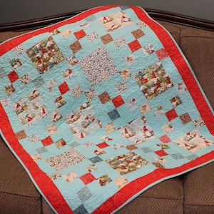 May include: A patchwork quilt featuring a light blue background with alternating squares of holiday-themed gnome prints, small floral patterns, and solid red squares. The quilt is finished with a wide, solid red border and intricate stitching.