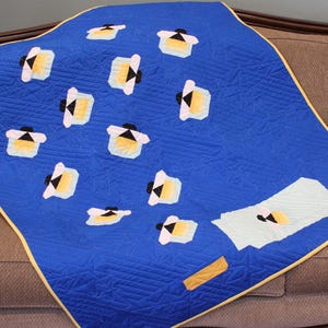 May include: A blue quilt with a repeating pattern of white, yellow, and black shapes. The quilt has a yellow border and is displayed on a brown sofa. The quilt's design is geometric and modern.