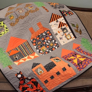 May include: A Halloween-themed quilt with a patchwork design featuring various houses, trees, and pumpkins. The quilt is made with a variety of fabrics in orange, black, and green.