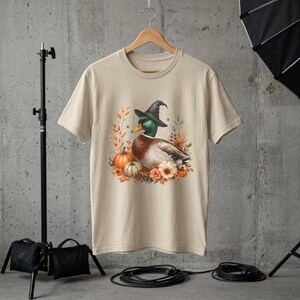 May include: A beige short-sleeve t-shirt featuring a graphic print of a mallard duck wearing a black witch hat. The duck is surrounded by orange pumpkins, autumn leaves, and flowers in shades of orange, brown, and cream on the front center.