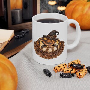 May include: A white ceramic mug featuring a graphic illustration of a round, brown, grumpy-looking toad wearing a dark brown witch hat, sitting in a pile of dirt. The mug has a standard handle and is filled with a dark liquid.