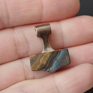 Könnte beinhalten: Ein Anhänger mit einem rechteckigen Tigerauge-Stein in braunen, goldenen und blaugrauen Streifen, gefasst in eine glatte, kupferfarbene Metallöse. Der Stein hat eine natürliche, unpolierte Textur mit sichtbaren Streifen auf der Oberfläche.