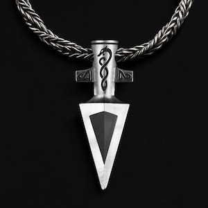 May include: A silver necklace with a pendant shaped like a spearhead. The spearhead has a black dragon design on the top and a black triangle in the center.