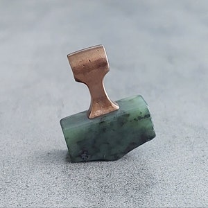 May include: A green stone pendant with a copper-colored bail. The stone is irregularly shaped and has a rough texture.