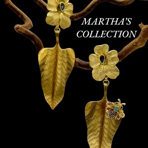 May include: Gold leaf earrings with a flower design and a small bee charm. The flowers have a blue gemstone in the centre. The earrings are hanging from a branch.